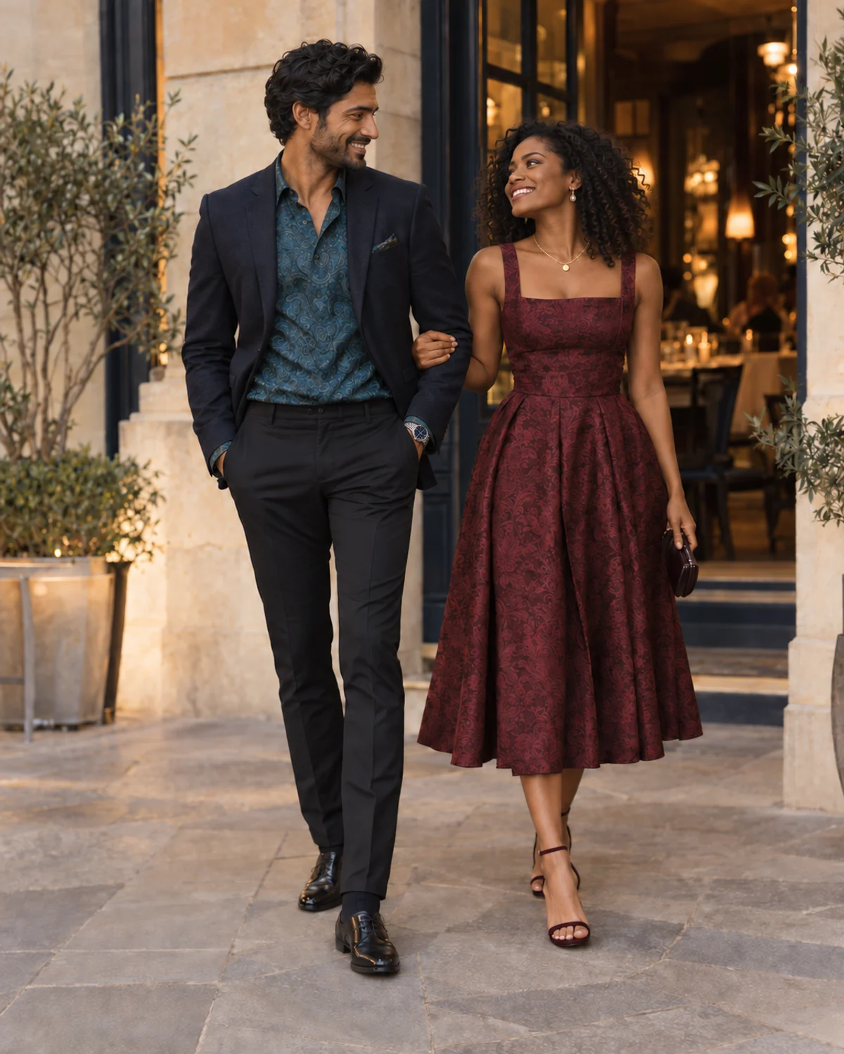 A couple in navy tailoring, a teal paisley shirt and visible wristwatch, and a burgundy jacquard dress with a gold necklace outside a restaurant